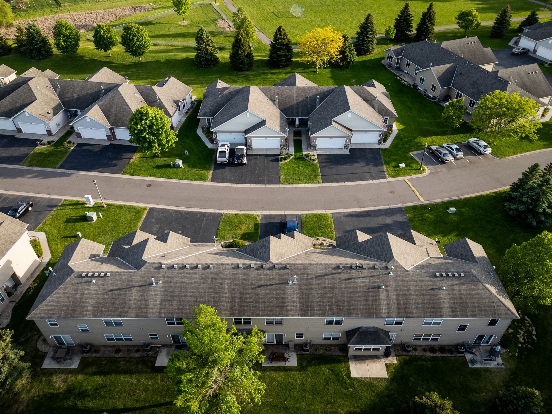 Aerial view of a suburban apartment complex with parked cars and trees along a curved road.