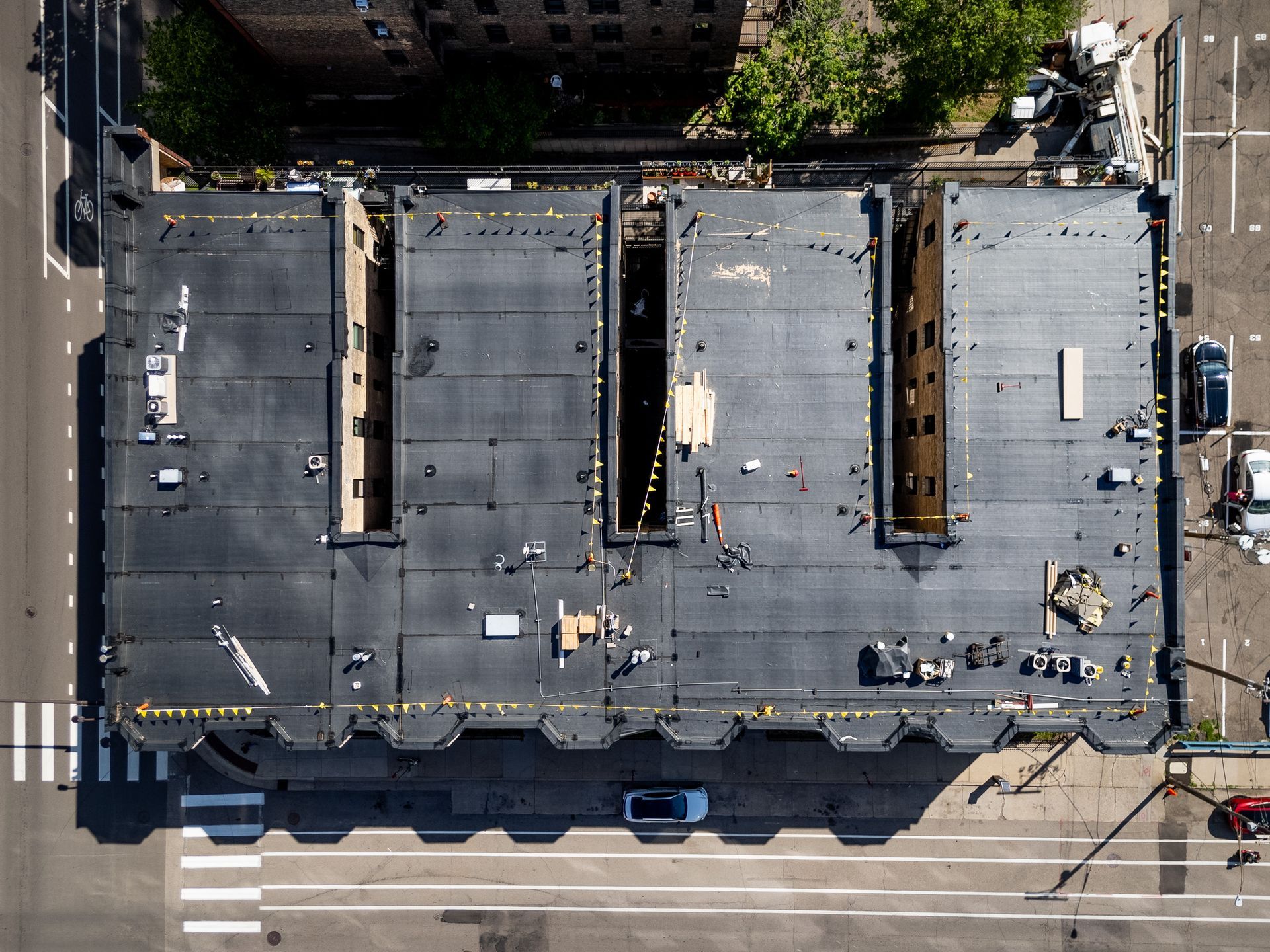 Aerial view of a flat-roofed city building with rooftop equipment, sidewalks, and parked cars nearby.