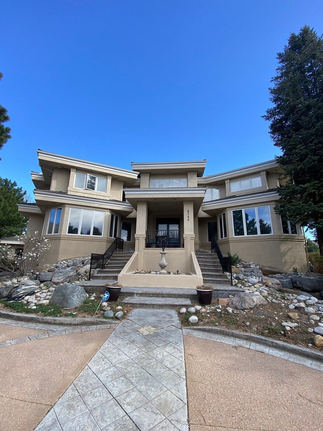 Stone mansion with central steps, glass windows, and a landscaped front yard under a clear blue sky