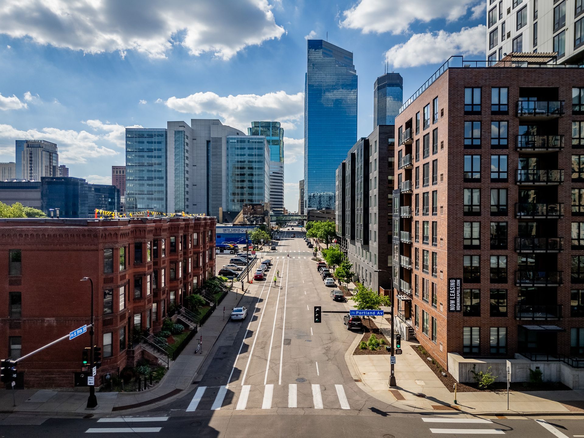 Downtown city street with tall modern buildings, a crosswalk, and blue sky with clouds