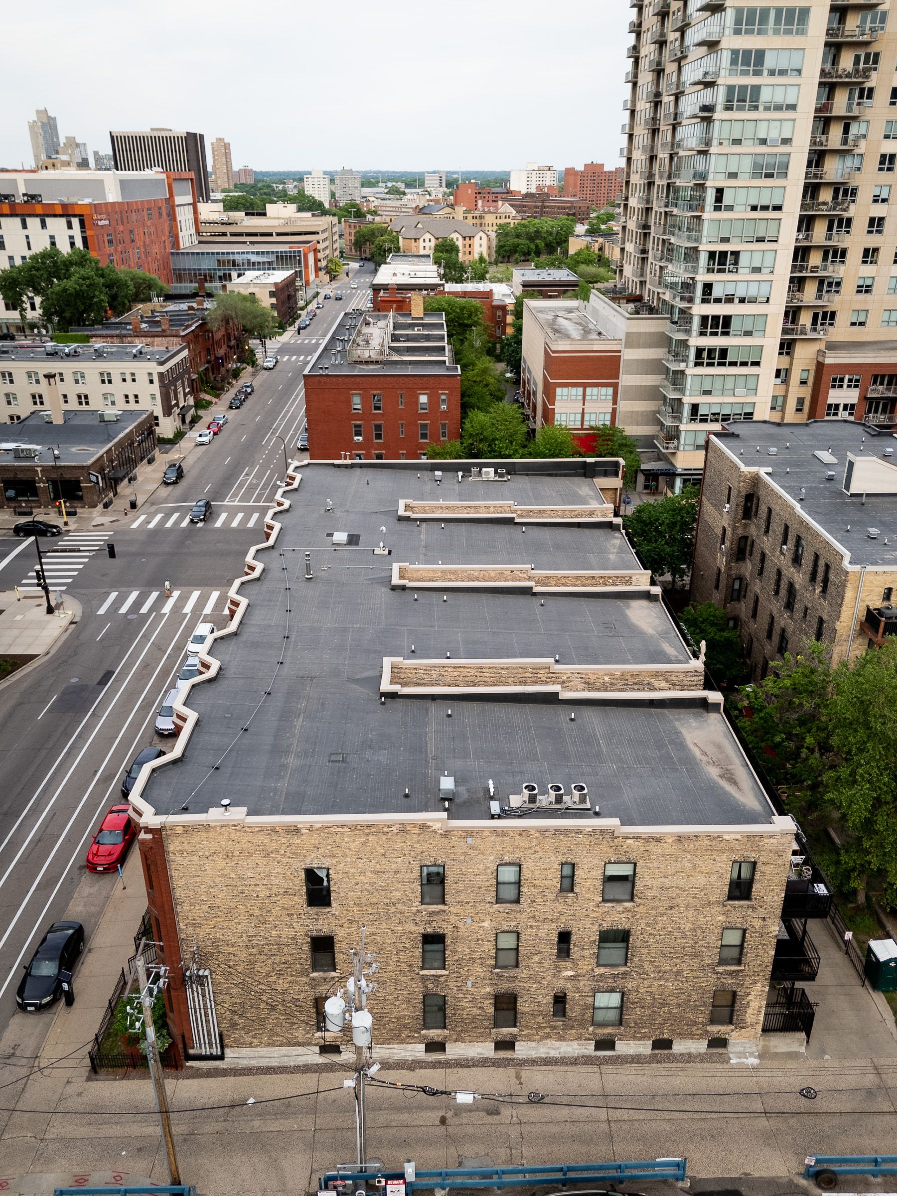 Aerial view of city street with brick buildings, parked cars, and a tall apartment tower under overcast sky