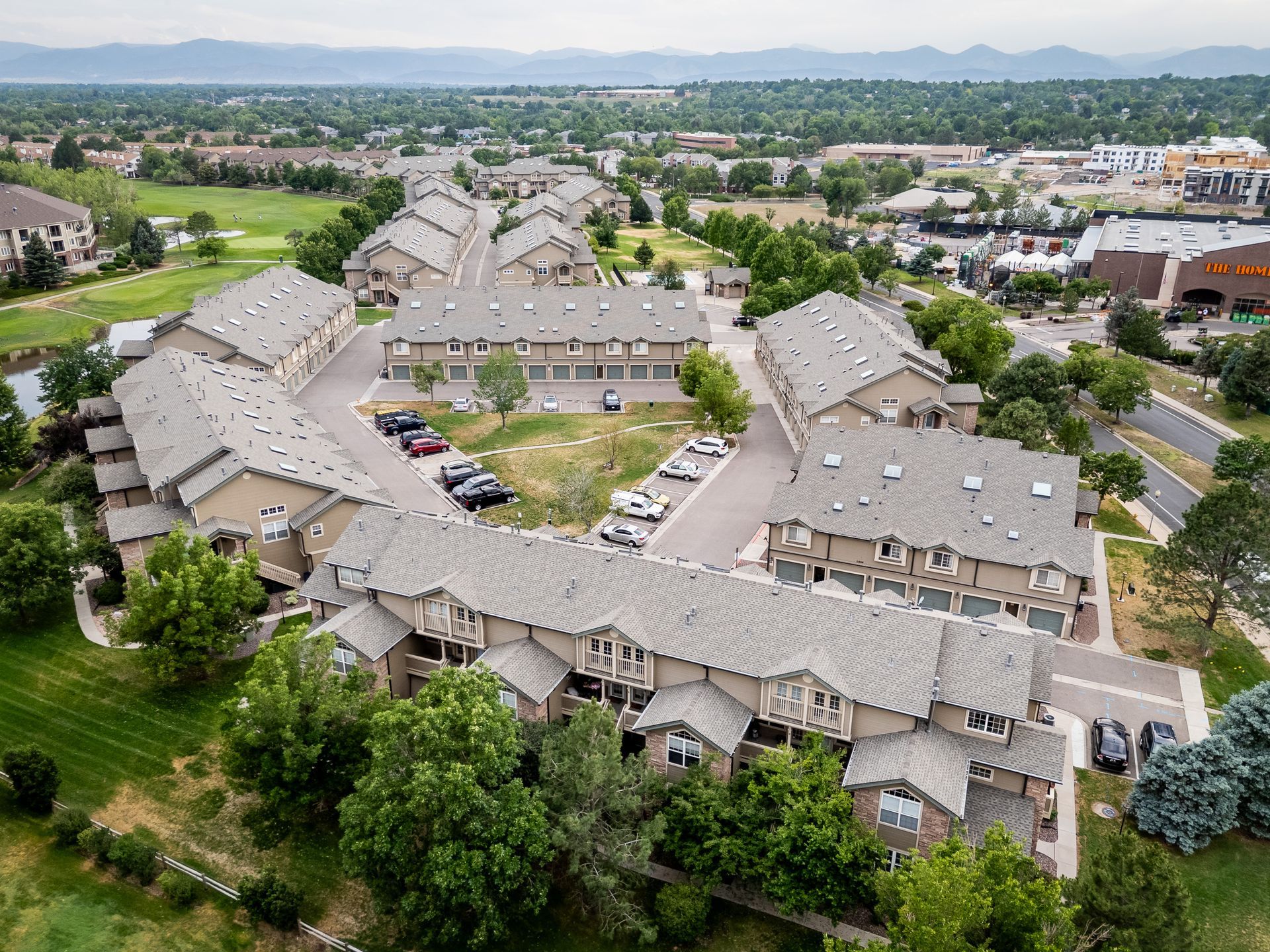 Aerial view of a large apartment complex with tan roofs, trees, parking lots, and a distant city skyline