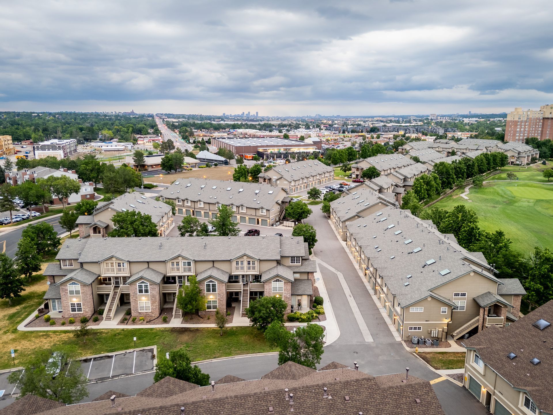 Aerial view of a suburban apartment complex with rows of gray rooftops and streets, overlooking a city skyline.