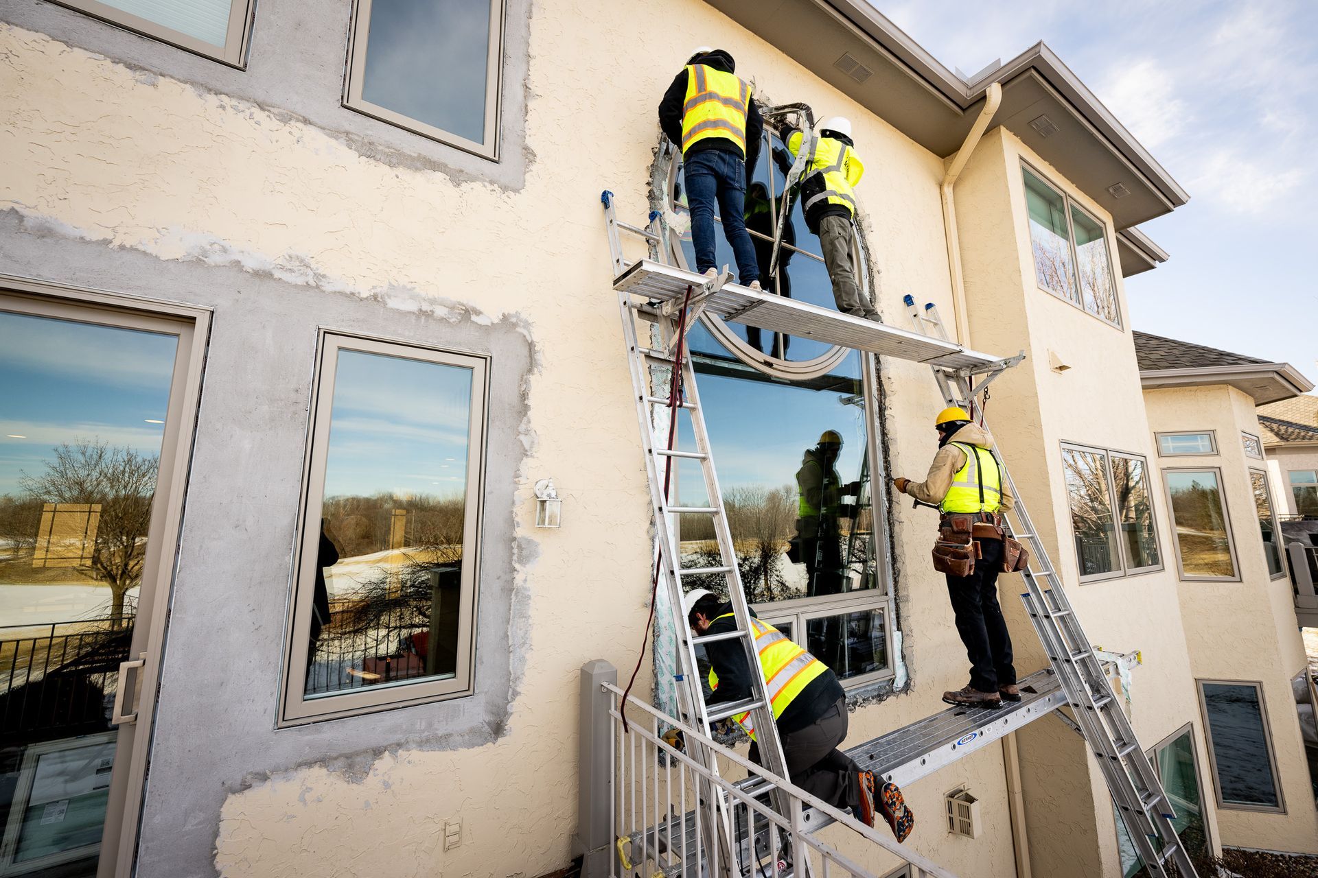 Workers in yellow vests repair a stucco building from metal scaffolding outside large windows