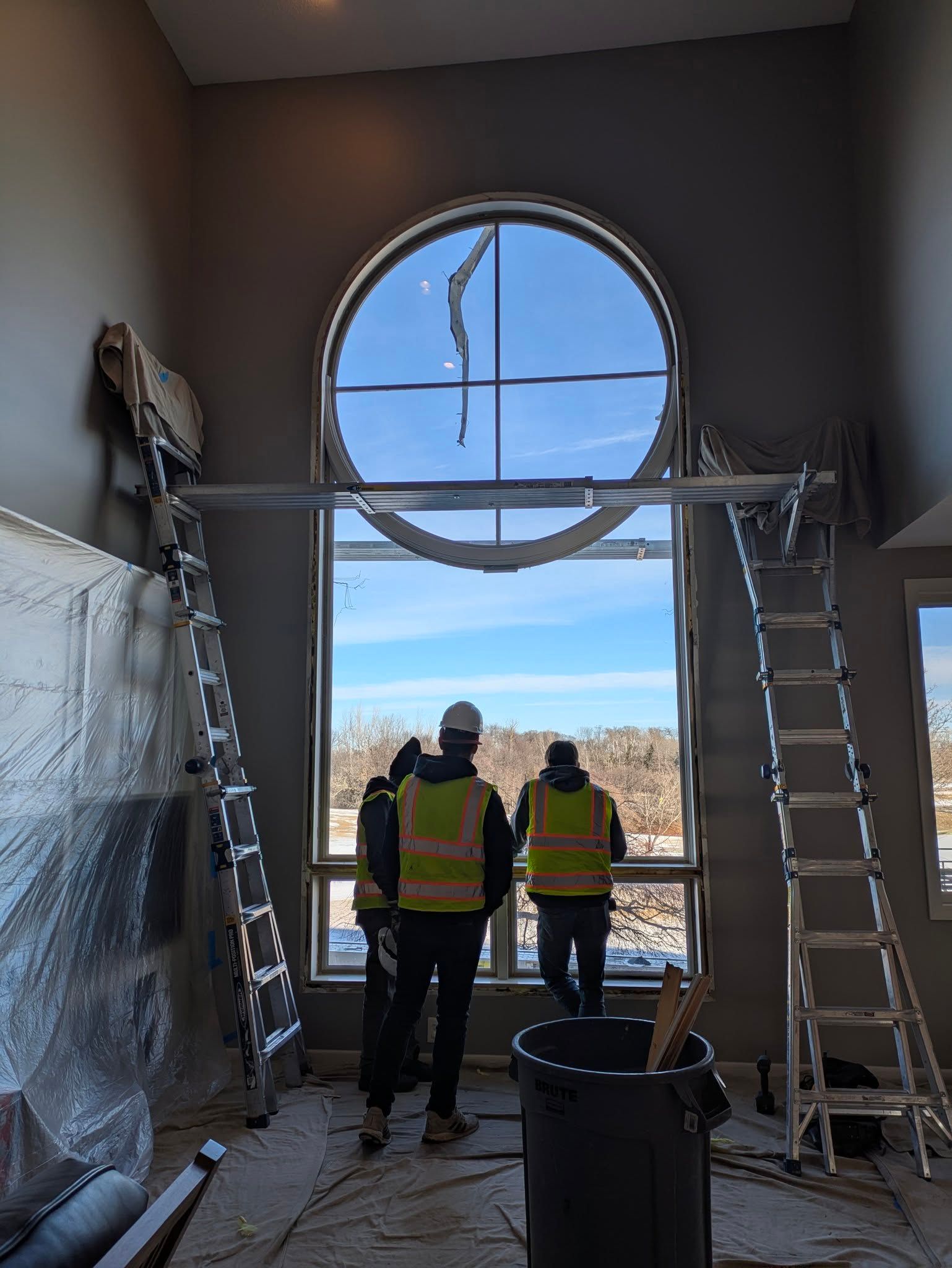 Two construction workers in safety vests stand by a large circular window in a room under renovation.