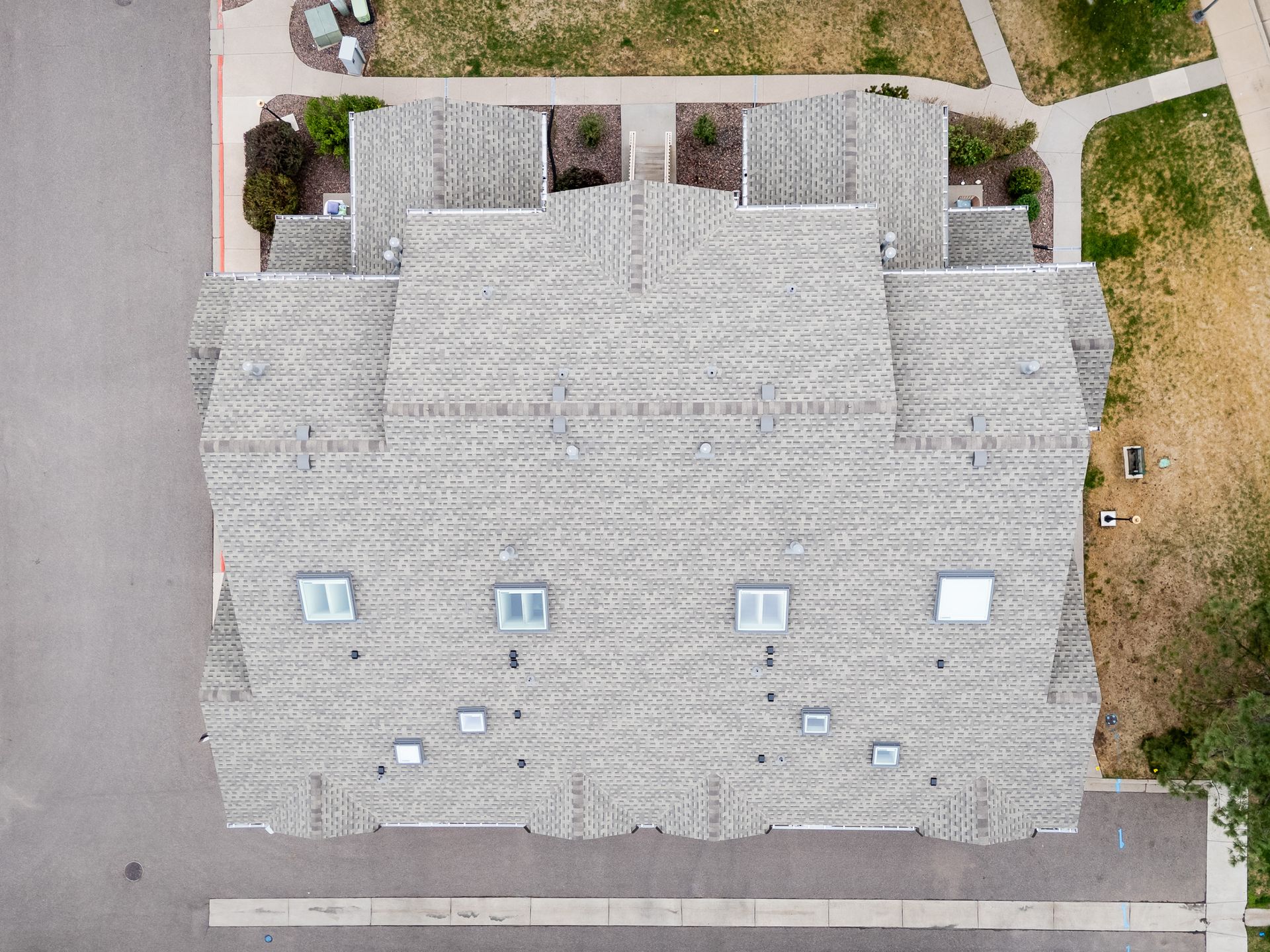 Aerial view of a gray-shingled house roof with several skylights beside a road and yard.
