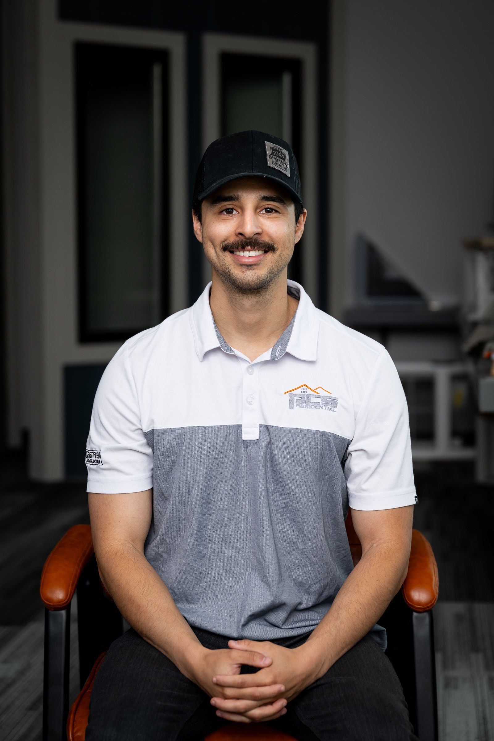 Man seated indoors, wearing a black cap and white-gray polo shirt, smiling at the camera.