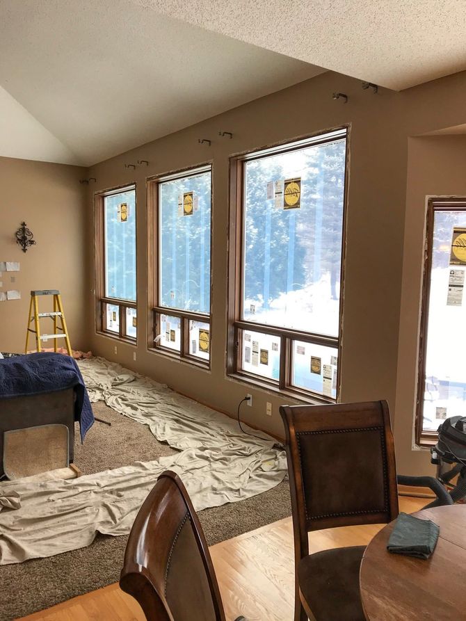 Sunlit living room with large windows, white rug, and wooden chairs near a dining table.