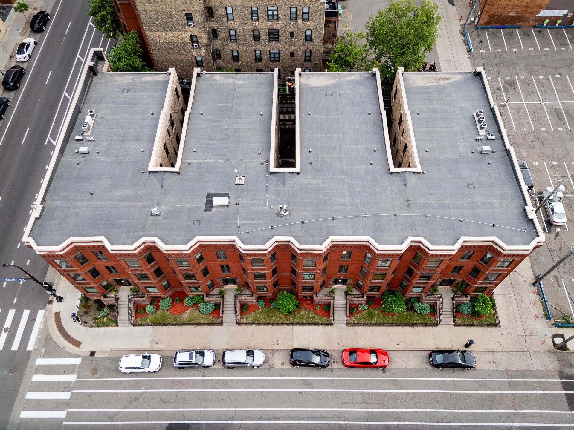 Aerial view of a red-brick apartment building with rooftop skylights, parked cars, and city streets below.