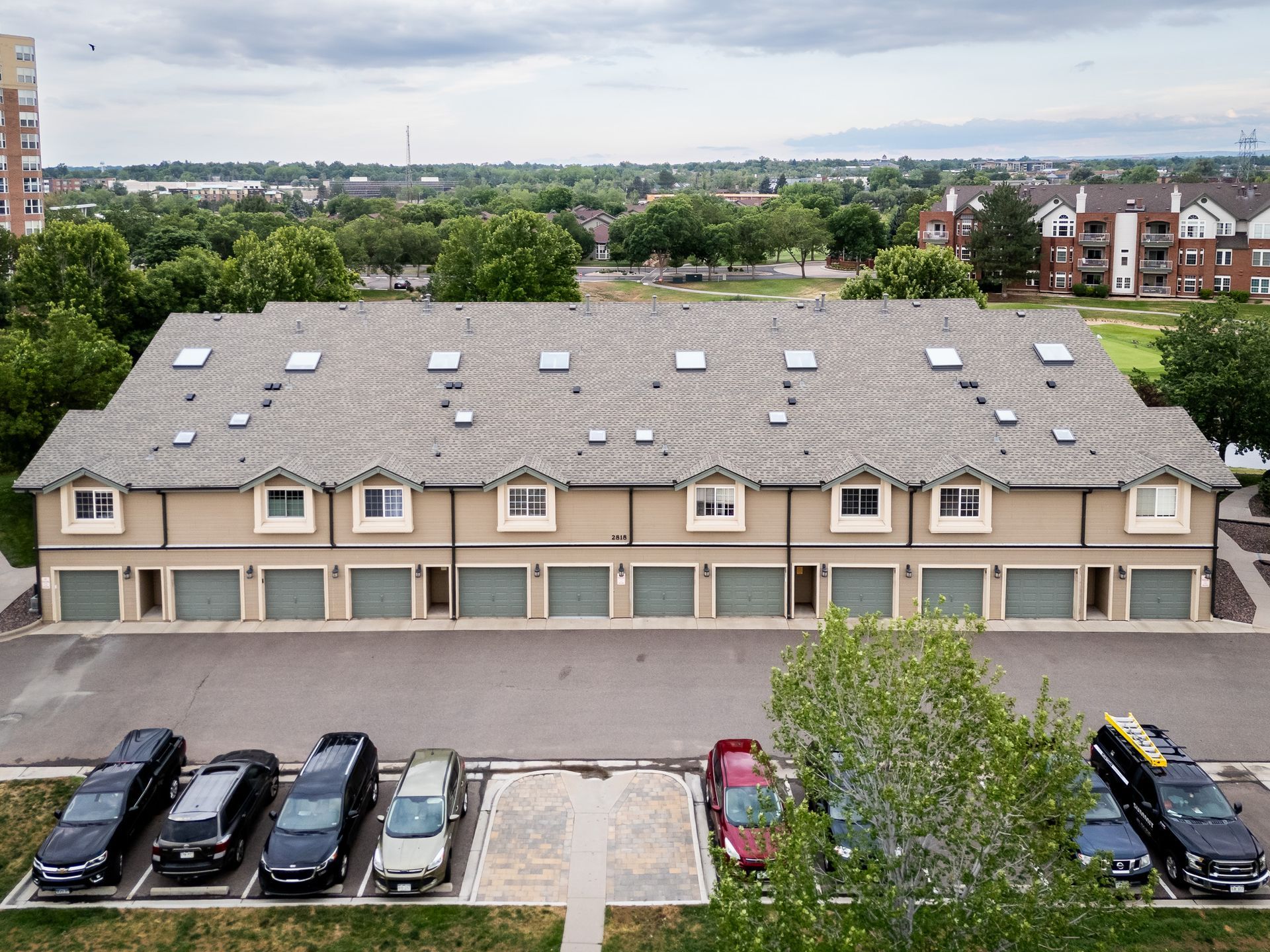 Aerial view of a beige apartment building with parked cars, trees, and a city skyline in the background