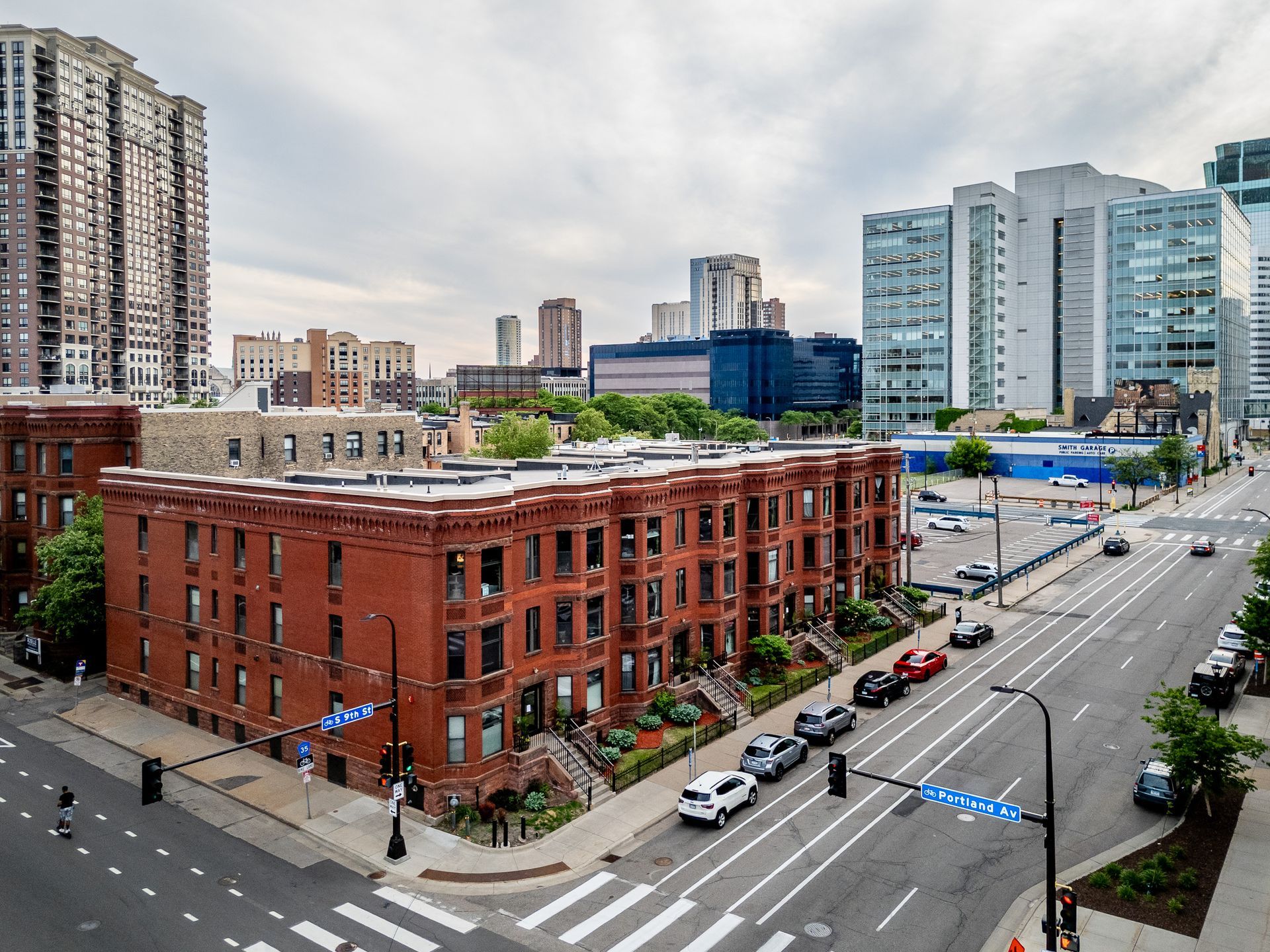City street with a red brick corner building, parked cars, and modern apartment towers under cloudy skies