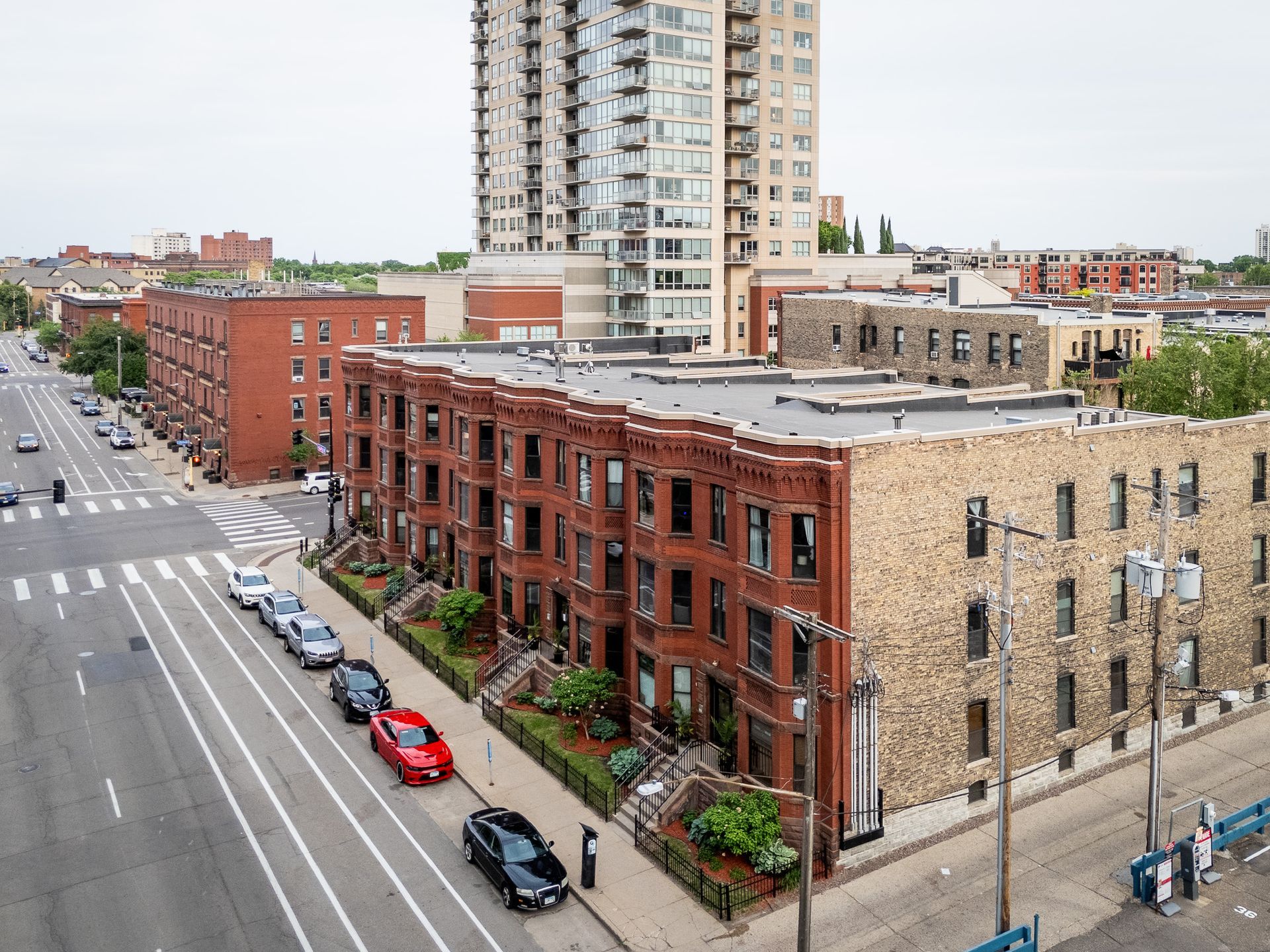 City street with red brick buildings, parked cars, and a tall apartment tower under a cloudy sky