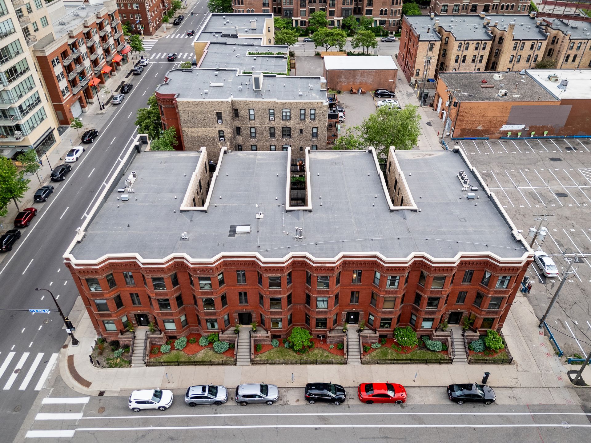 Aerial view of a red-brick apartment building beside a city street with parked cars and nearby buildings