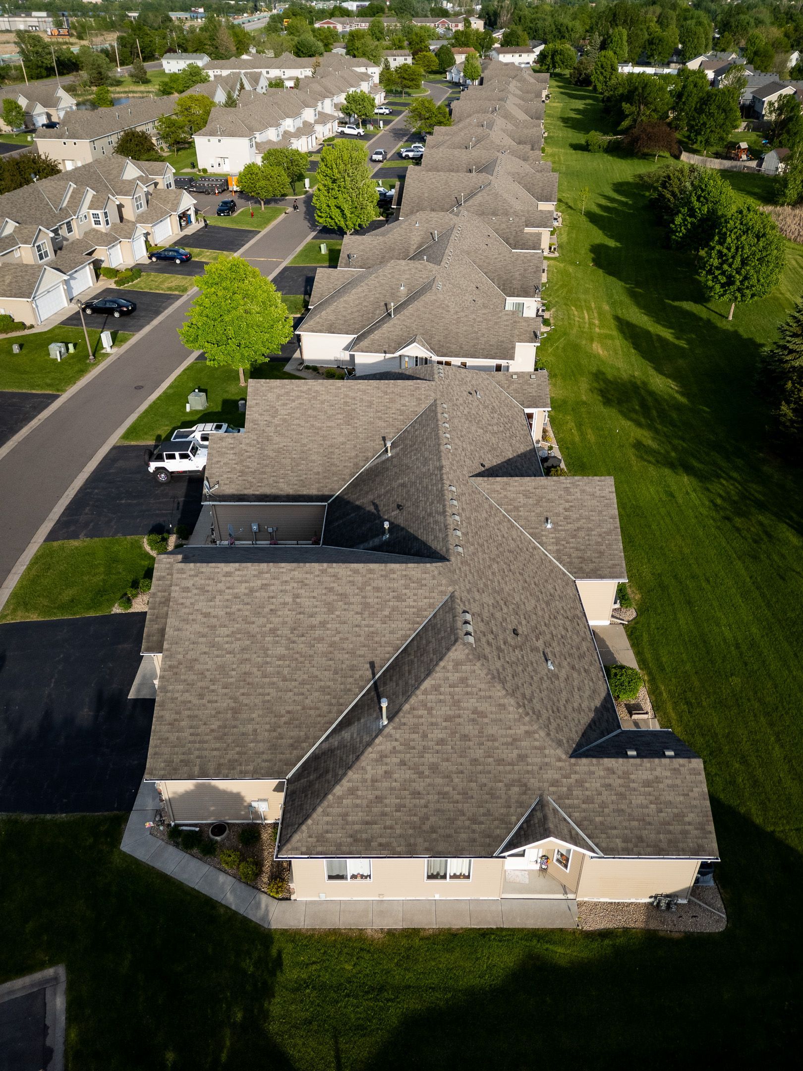 Aerial view of rows of suburban houses with gray roofs and a green wooded area alongside.