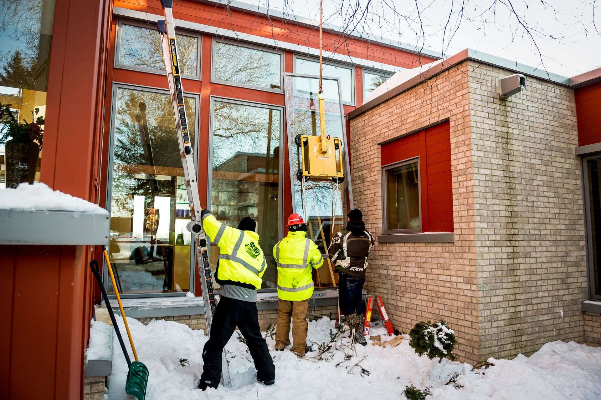 Workers in yellow vests inspect a building site beside a brick structure in snow.