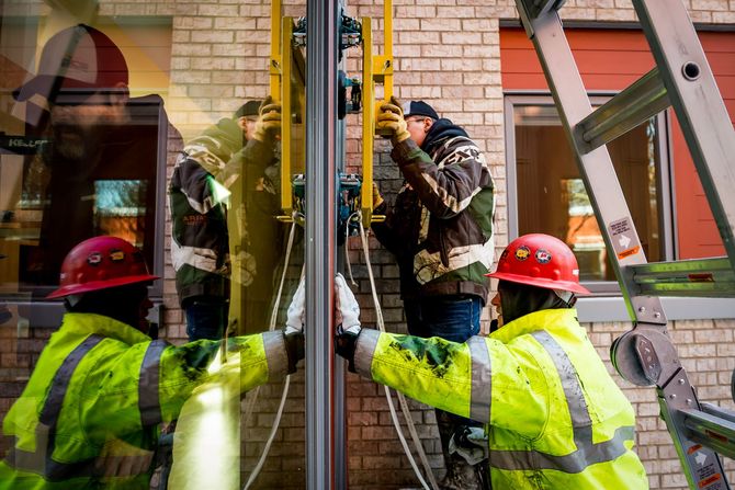 Firefighters in neon jackets and red helmets work by a glass doorway beside a ladder truck.