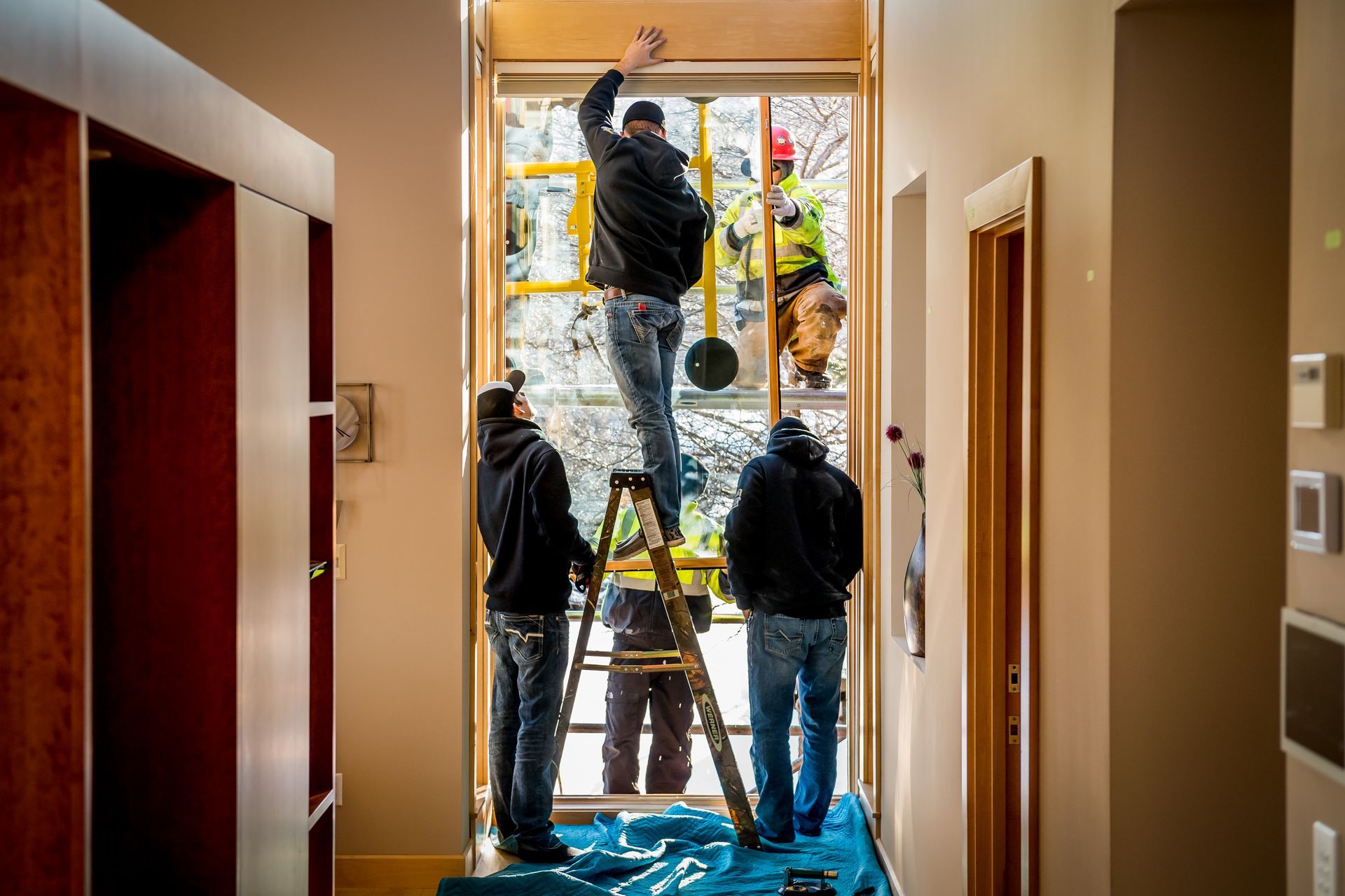 Workers installing a large window frame in a hallway, with ladders, tools, and protective plastic on the floor.