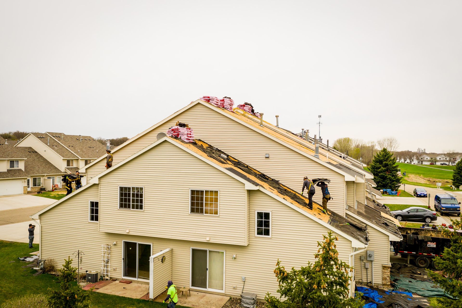 Workers repairing a beige house roof with pink shingles and ladders in a suburban neighborhood