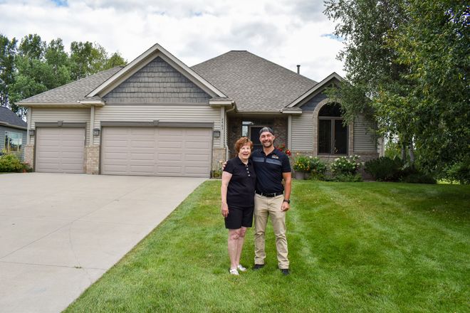 Two people standing on a lawn in front of a beige suburban house with a three-car garage.