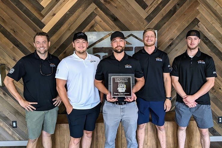 Five men in black and white polos hold a plaque in front of a wood chevron wall.
