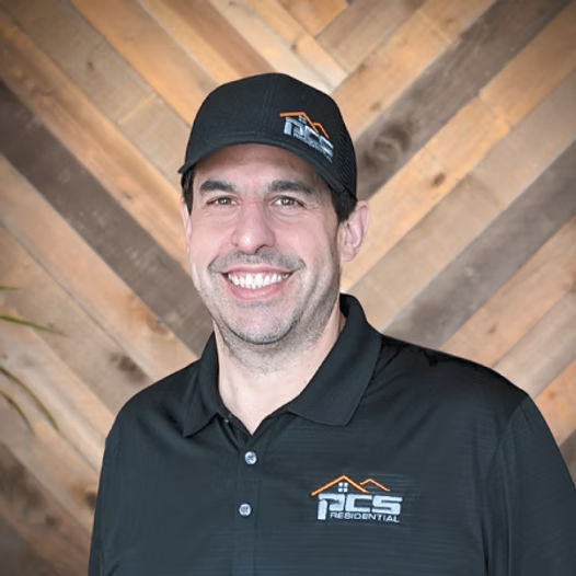 Smiling person in a black cap and polo shirt with a logo, standing before a wood plank wall