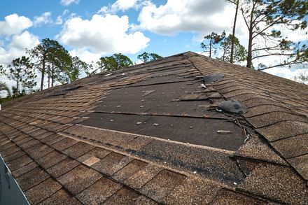 Damaged shingle roof with missing patches under a partly cloudy sky