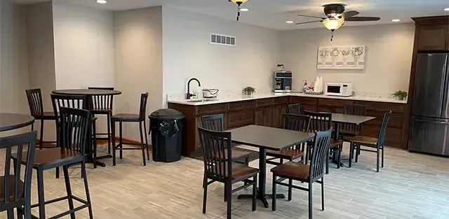 Empty kitchenette with barstools, tables, sink, and cabinets in a carpeted common area.