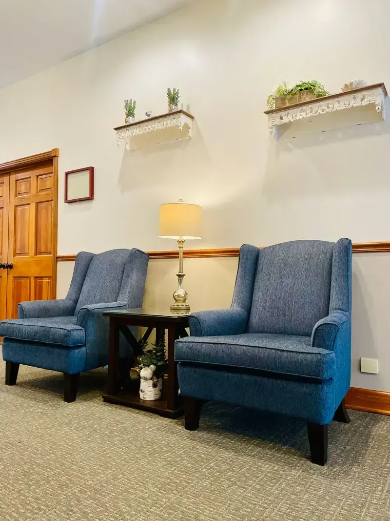 Waiting area with two blue chairs, small table with lamp, wall shelves, and wooden door on beige walls.