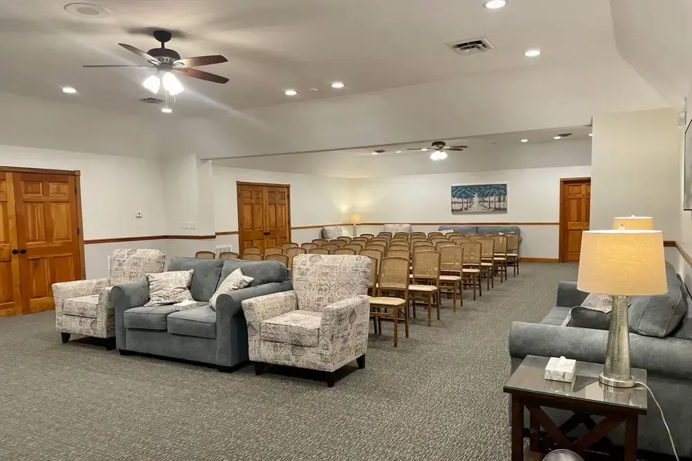 Spacious meeting room with rows of chairs, gray sofas, ceiling fans, and a table lamp.