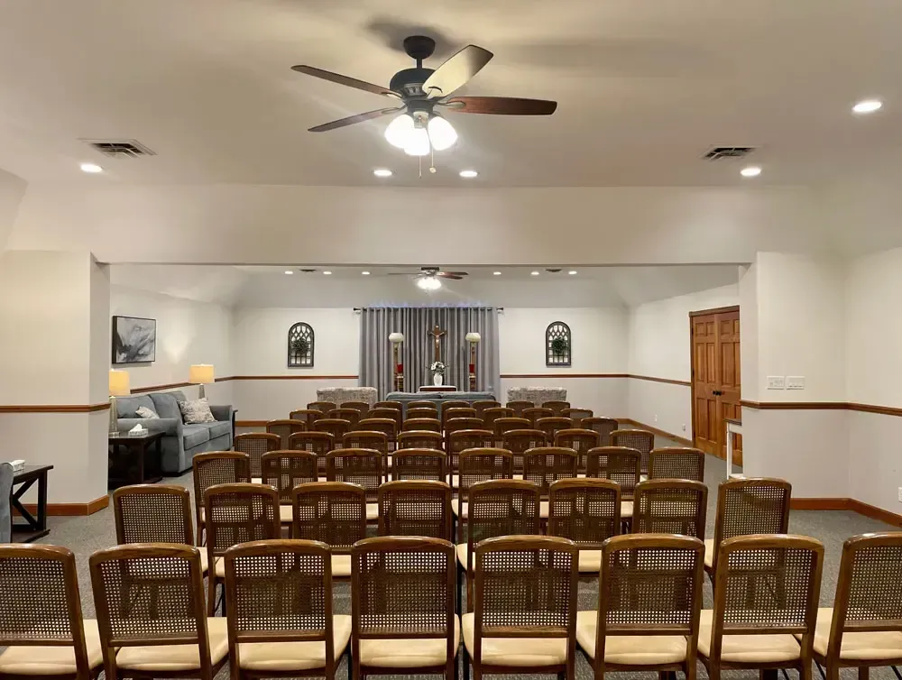 Empty church hall with rows of wooden chairs facing an altar and ceiling fans.