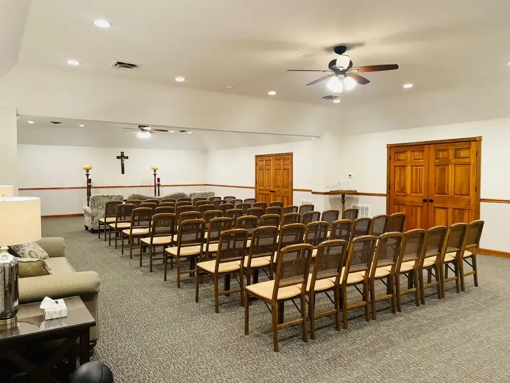 Empty chapel room with rows of wooden chairs, white walls, ceiling fans, and a cross at the front.