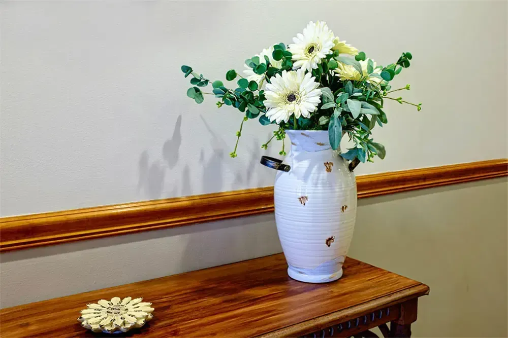 White vase of daisies and greenery on a wooden table against a beige wall