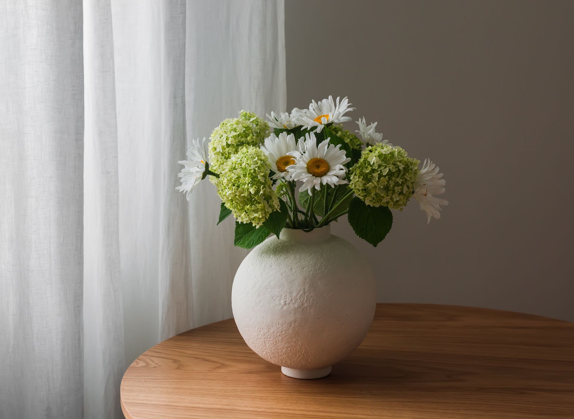 White vase with white daisies and green flowers on a round wooden table by a sheer curtain.