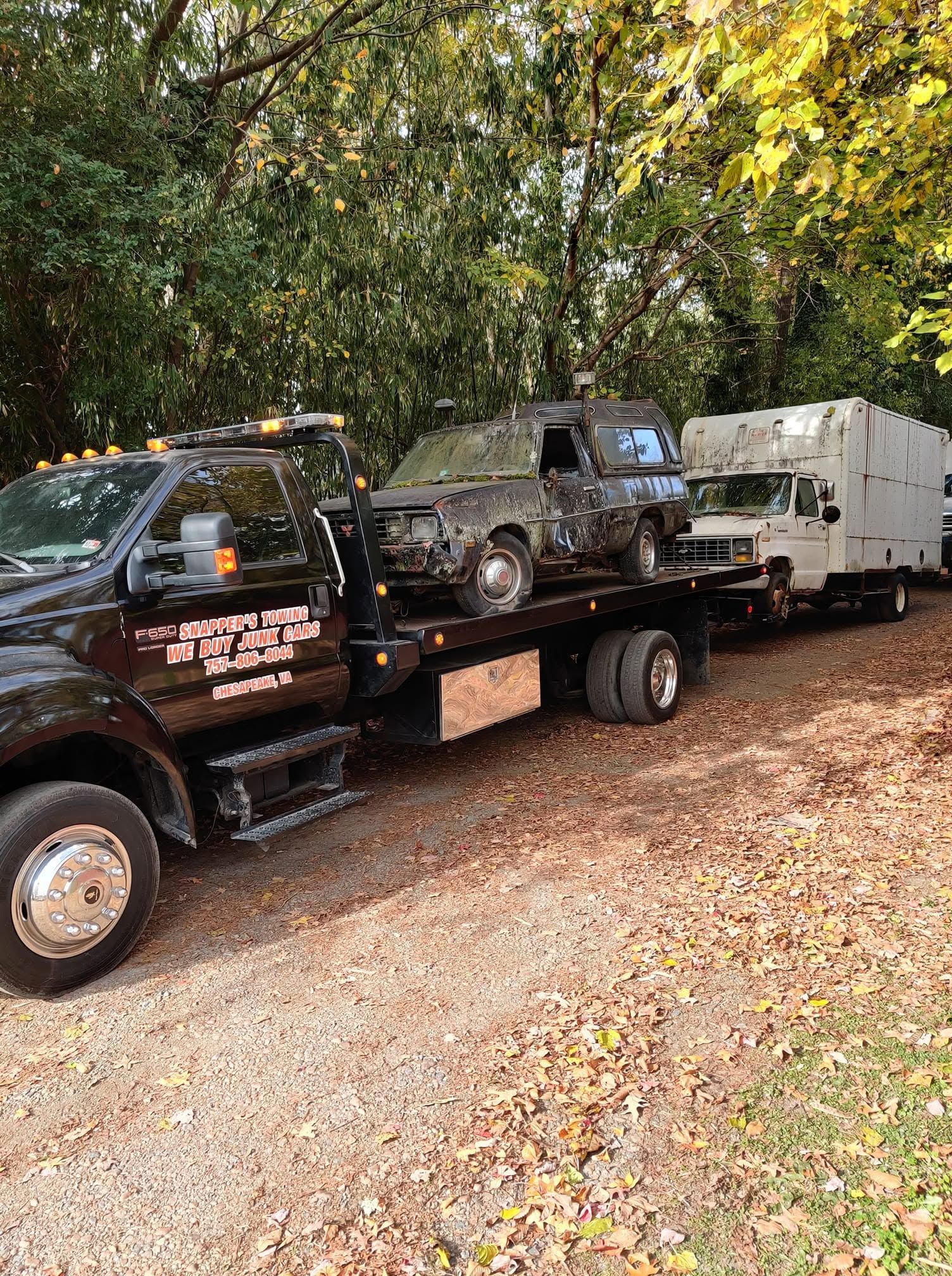 Tow truck hauling a burned-out SUV and a white trailer in a wooded area