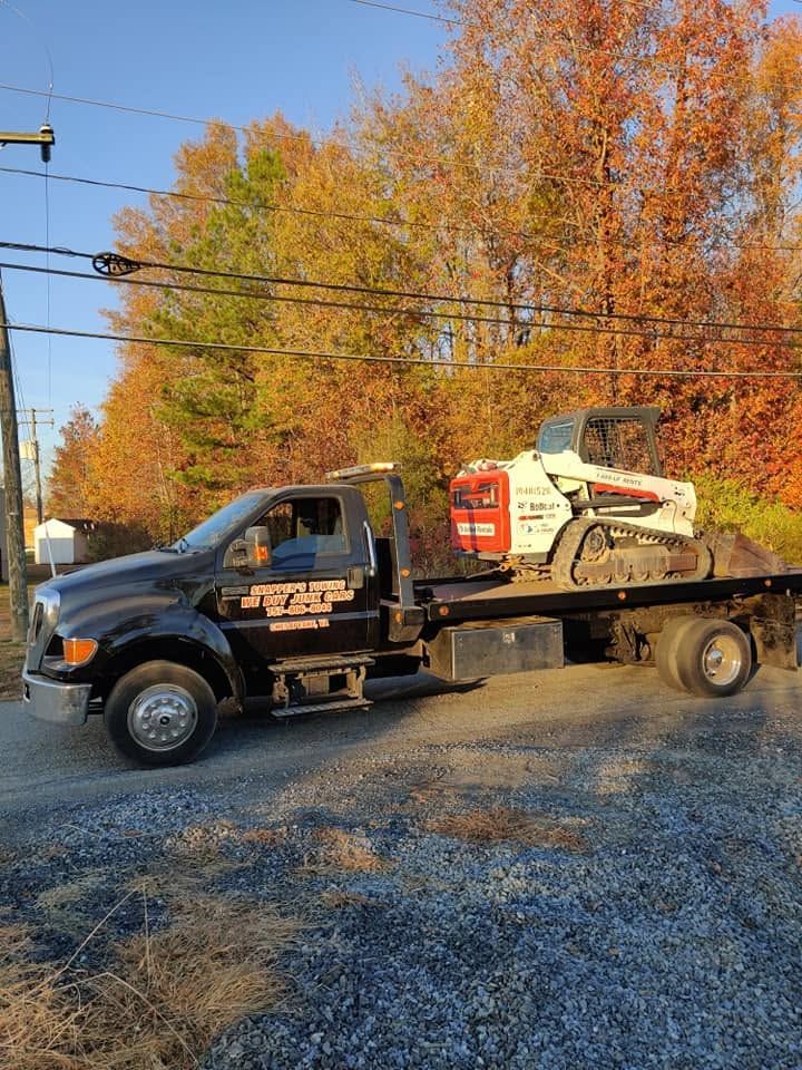 Tow truck carrying a white skid steer on a flatbed, parked on a gravel road with autumn trees behind.