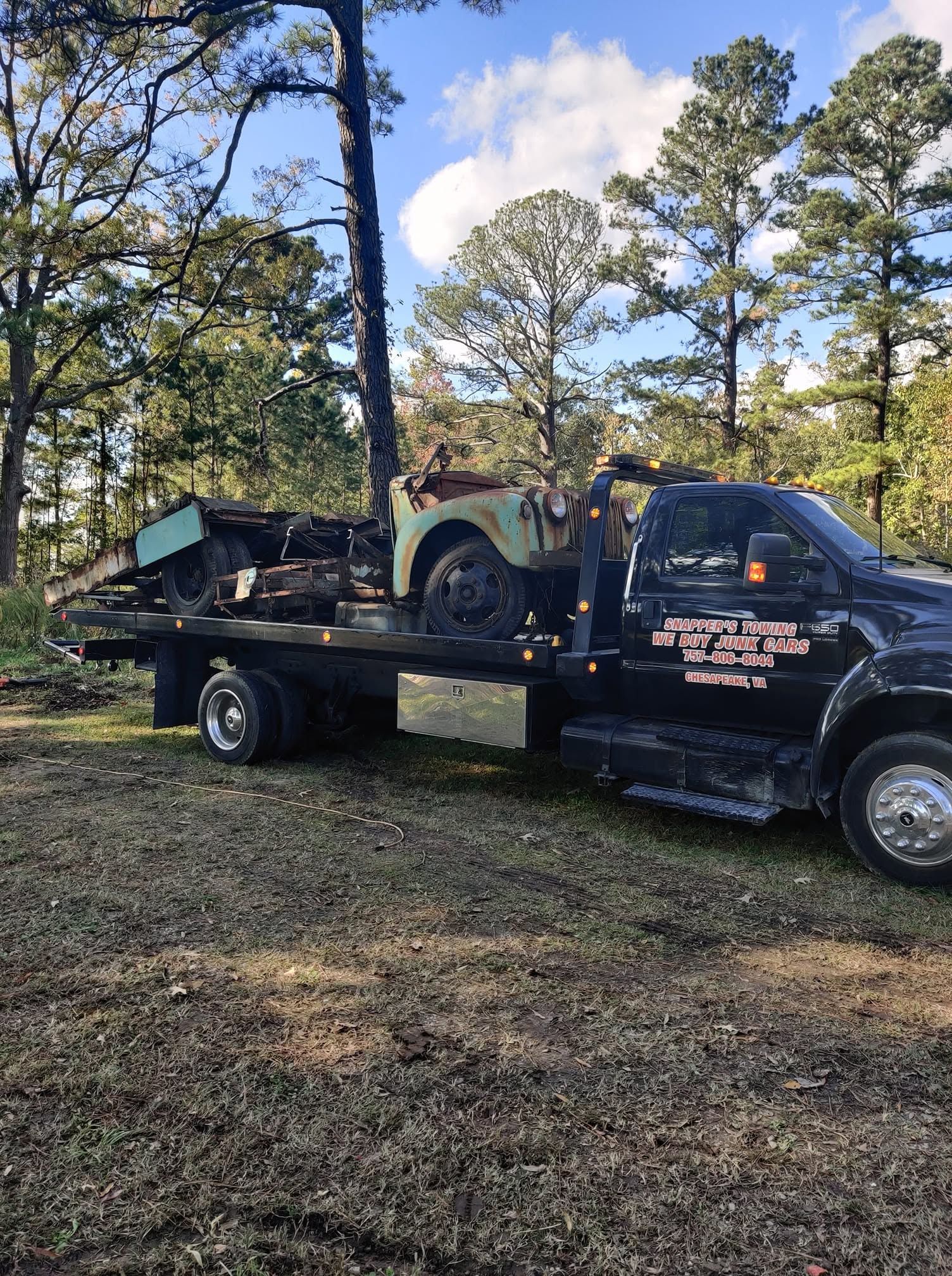 Tow truck hauling a green vintage vehicle on a flatbed in a wooded area