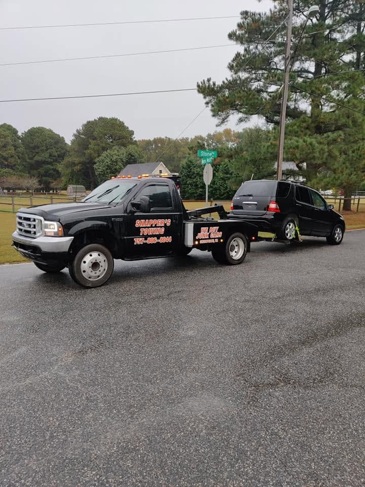 Black tow truck hauling a black sedan on a wet roadside under overcast skies.
