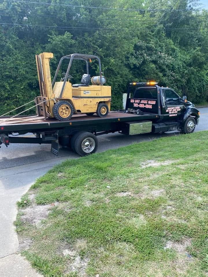 Flatbed tow truck carrying a yellow forklift on a suburban driveway with trees in the background