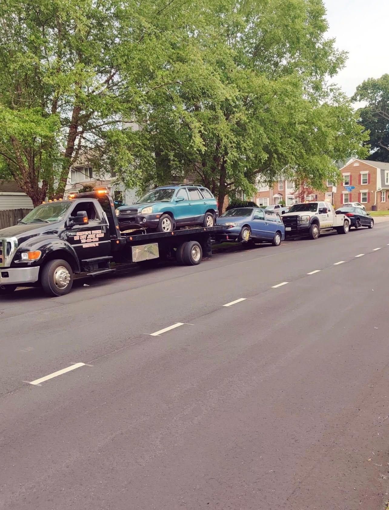 Tow truck hauling several parked cars along a tree-lined street beside brick houses