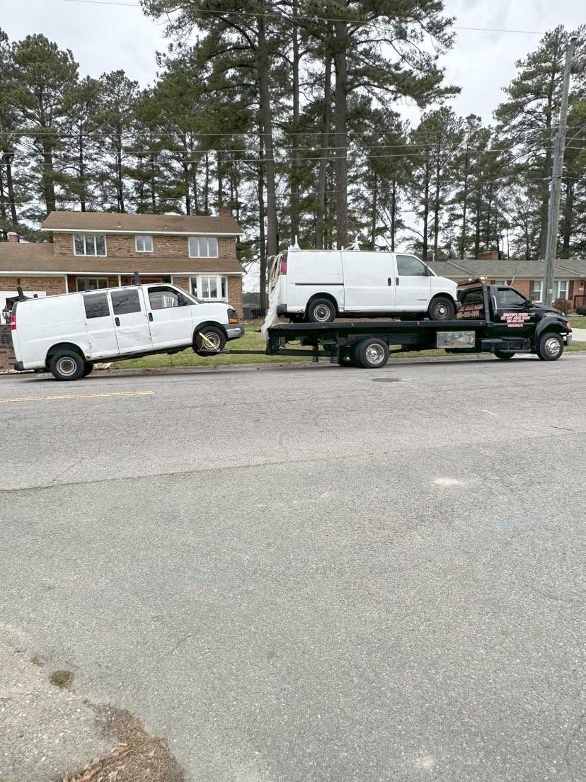 White pickup and utility truck on a flatbed trailer parked in front of a house with pine trees.