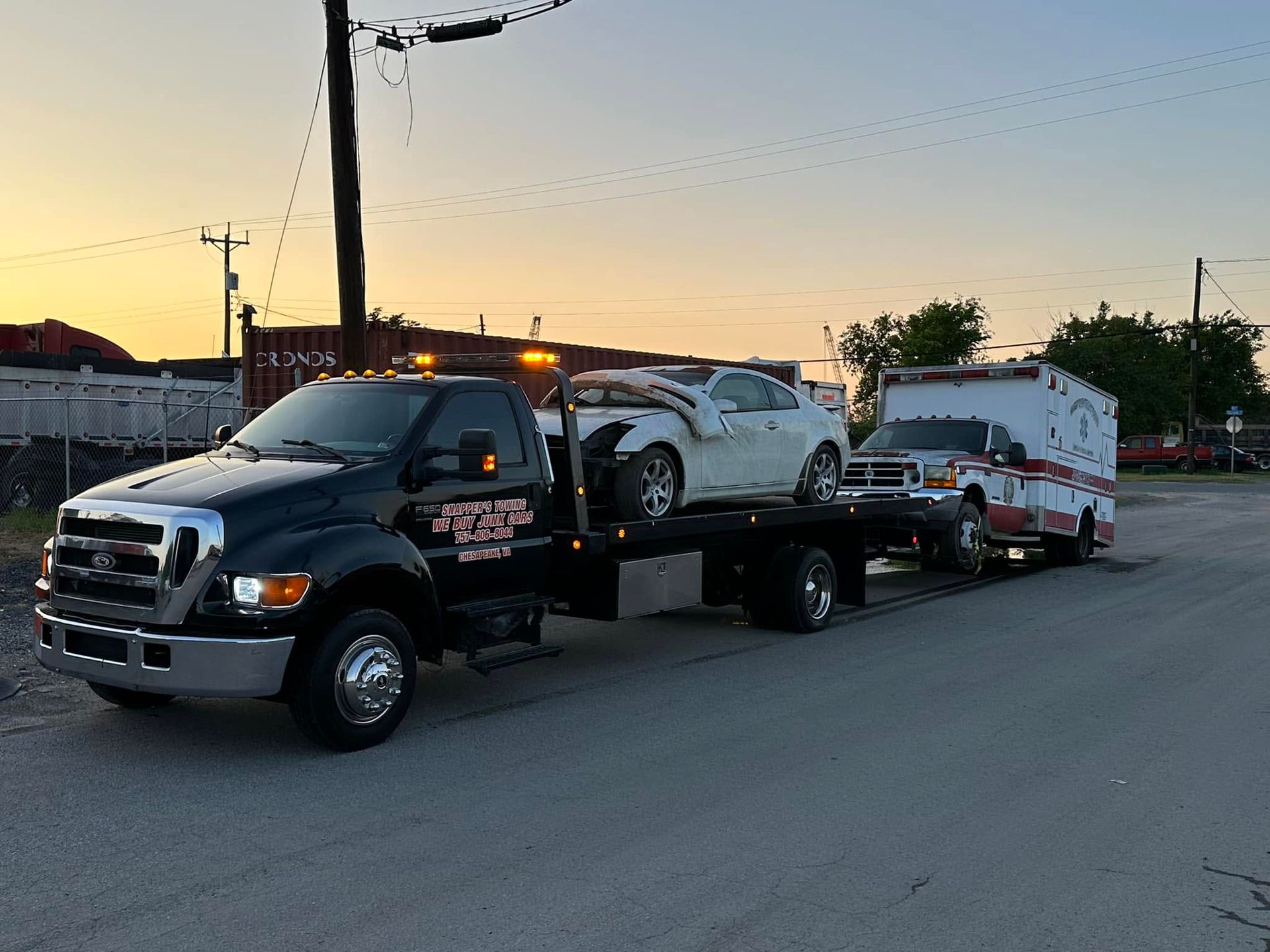 Tow truck hauling two wrecked cars on a road at sunset