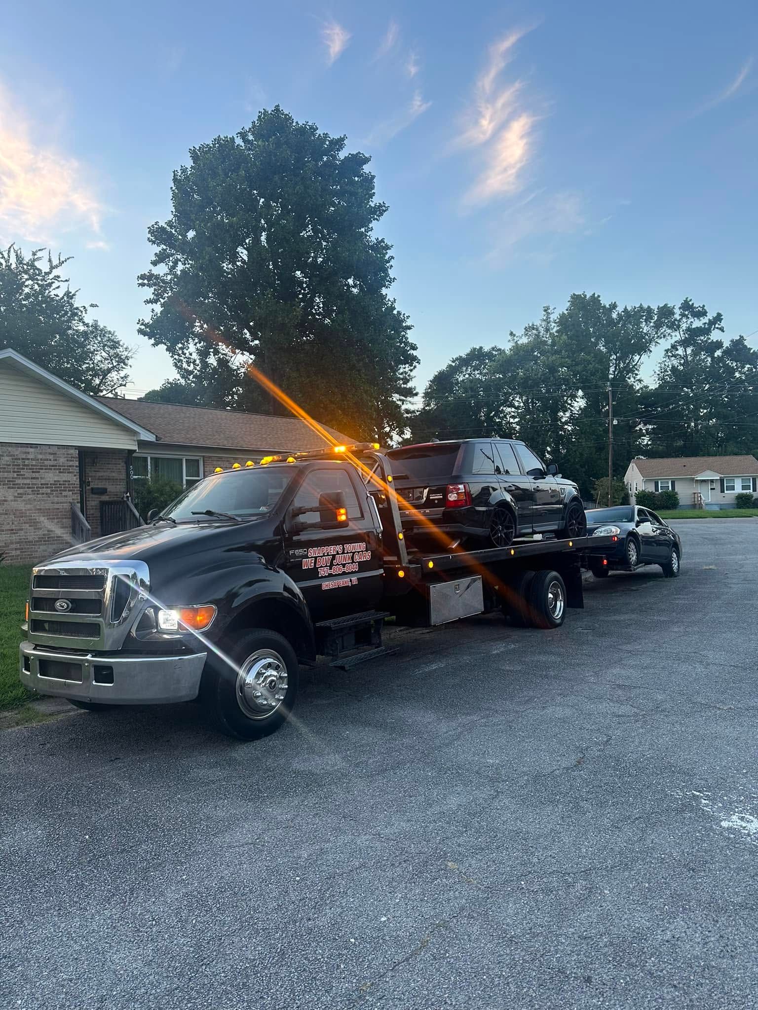 Tow truck carrying a white vehicle on a gravel lot at dusk, with flashing amber lights.