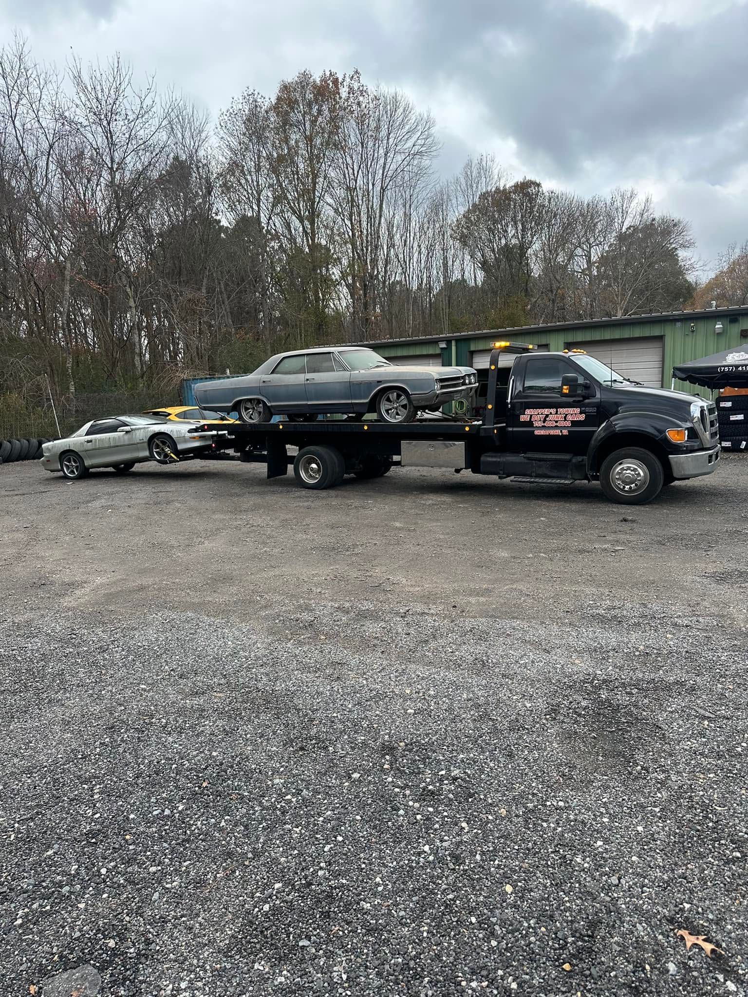 Tow truck hauling a black SUV in a gravel lot beside parked cars and trees