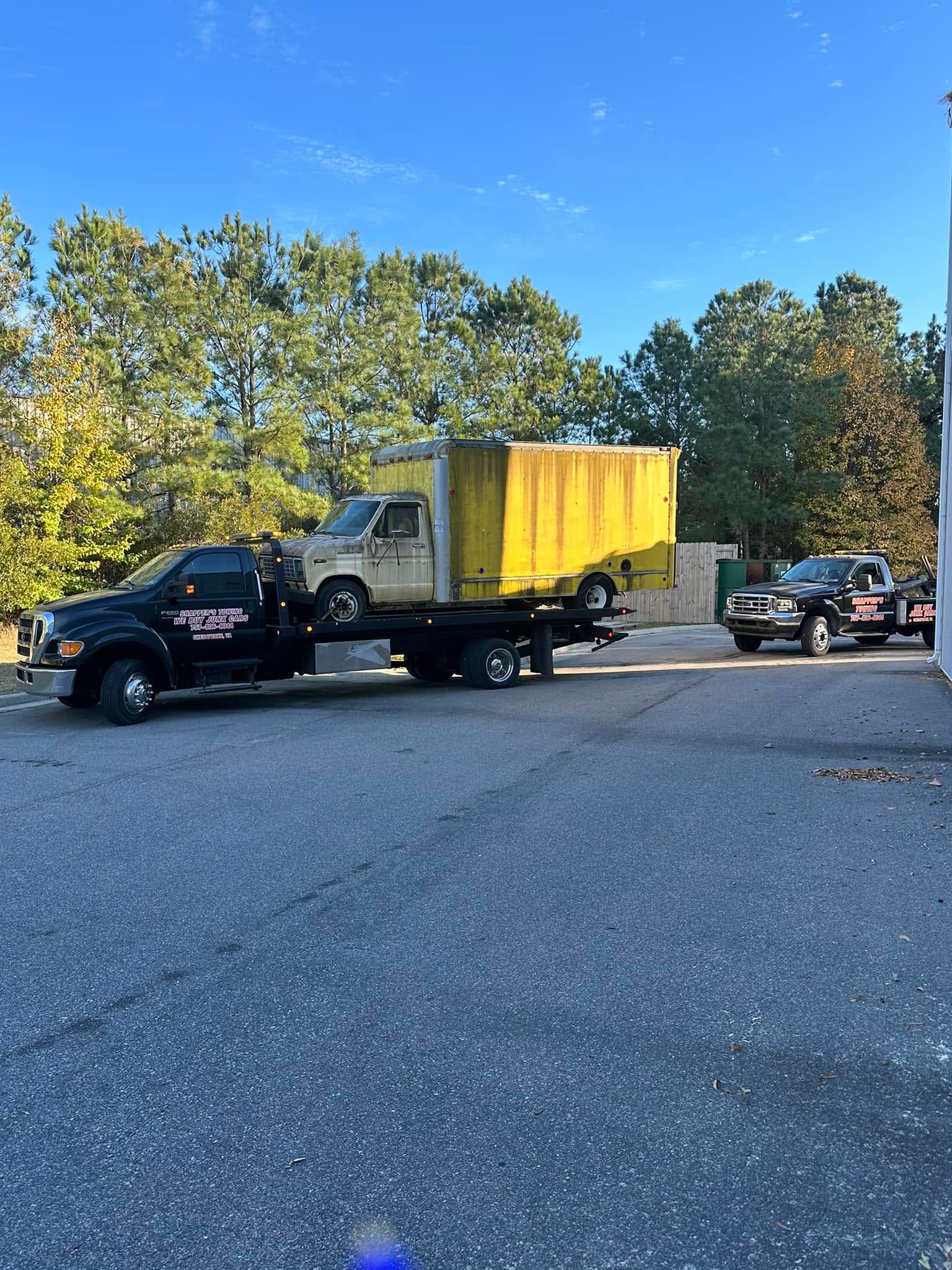 Tow truck hauling a yellow dumpster trailer on a road with trees and parked cars.