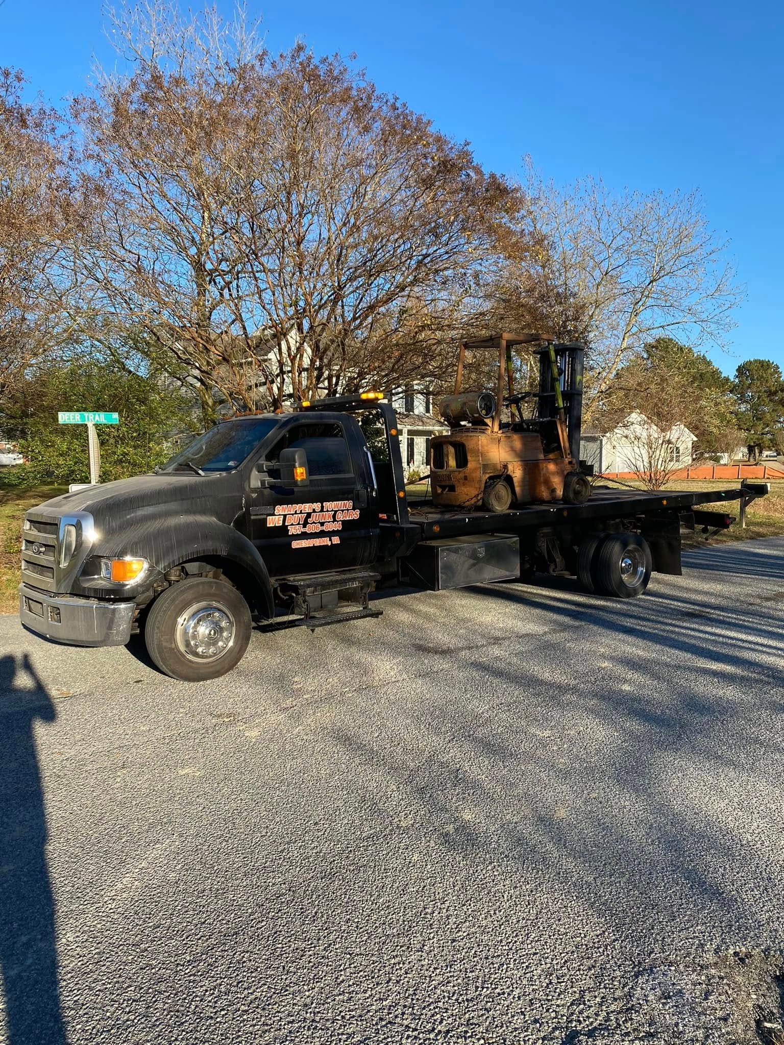 Black tow truck carrying a yellow bulldozer on a gravel lot under bare trees and blue sky