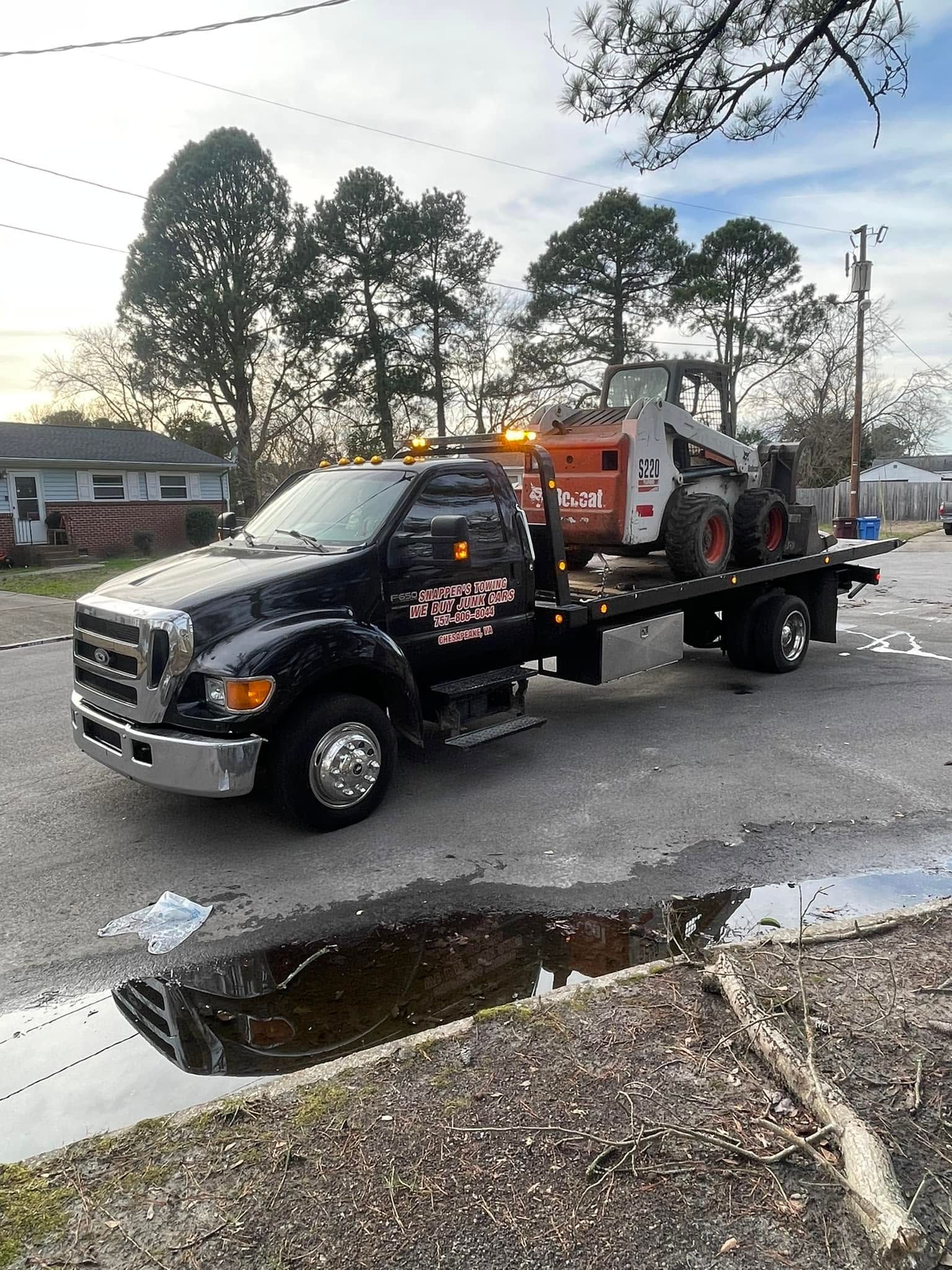Black tow truck with amber lights parked on a wet street near a puddle and trees