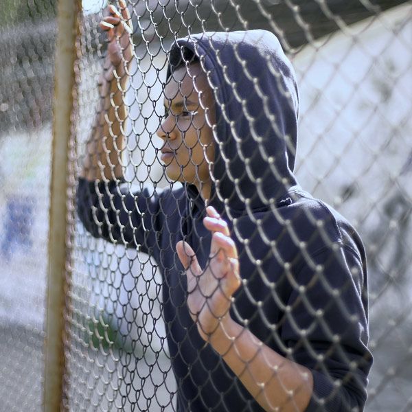 Person in a black hoodie standing behind a chain-link fence, looking through it with hands raised.