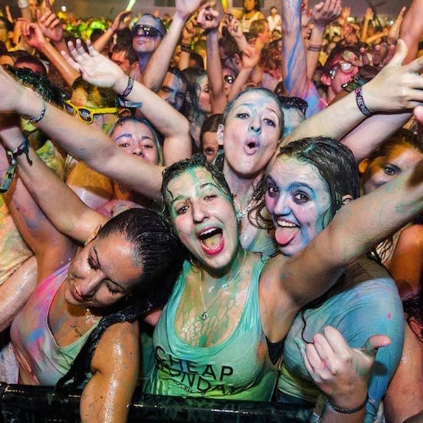 Crowd at a colorful festival, smiling and cheering with arms raised and faces covered in paint