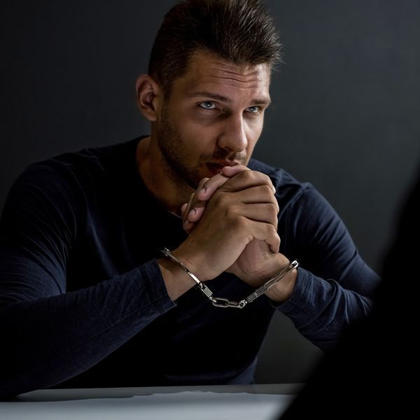 Handcuffed man in a dark shirt sits at a table, hands clasped in thought under dim light