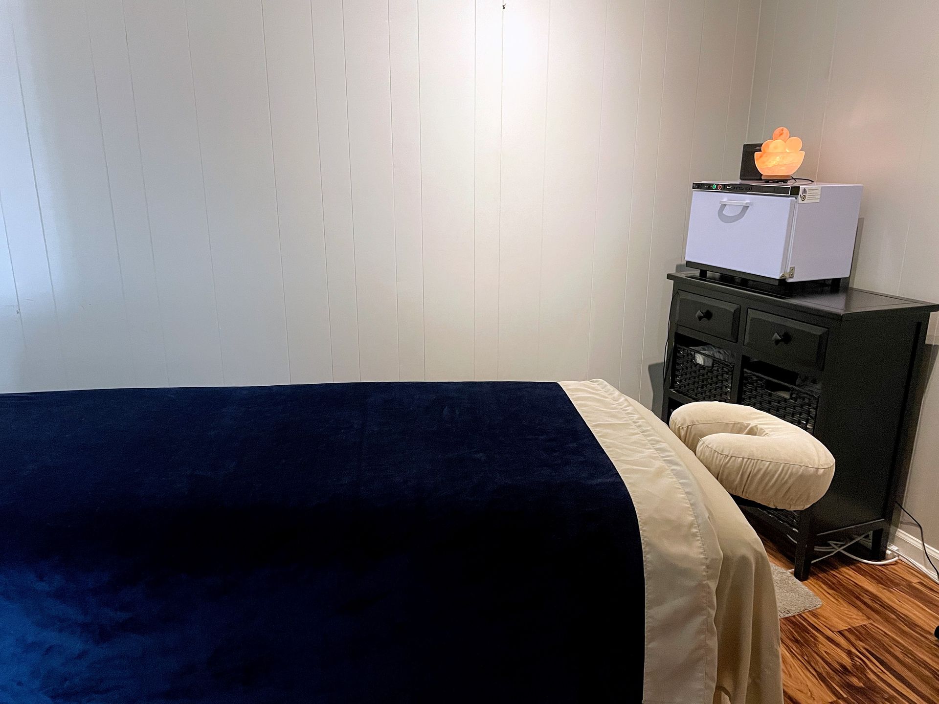 Bedroom with a dark blue bedspread, dresser, white bin, and small orange lamp near a window.